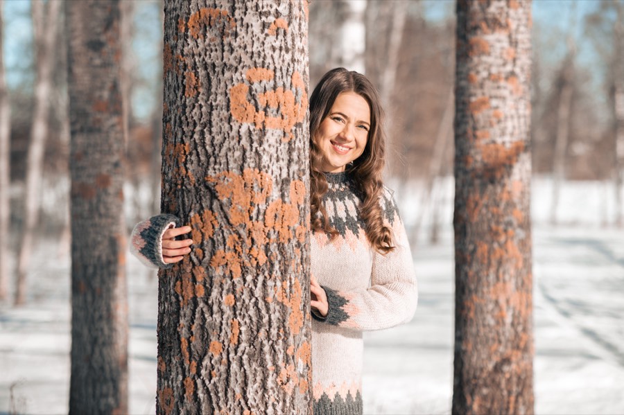 Henkilö hymyilee puun takaa talvisessa metsässä